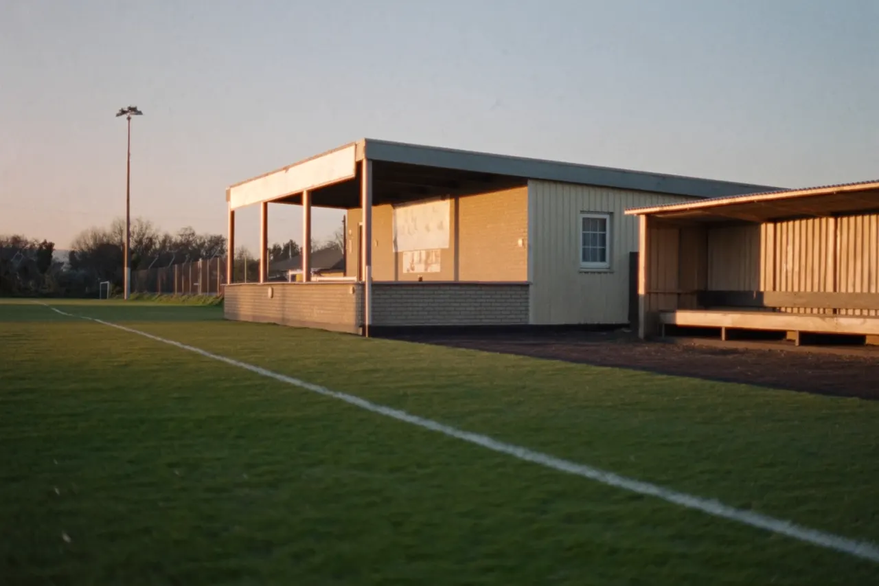 A suburban football clubhouse and covered players' dugout beside a grass pitch in late afternoon sun